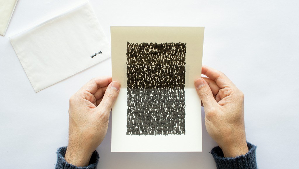 Hands holding a cream sheet of dense black marks, with a blank envelope beside it.